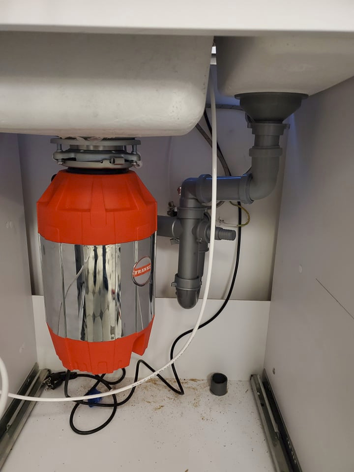 Zamontowany rozdrabniacz do odpadów Franke pod zlewem dwukomorowym, widoczne szare rury odpływowe i biały przewód doprowadzający wodę.