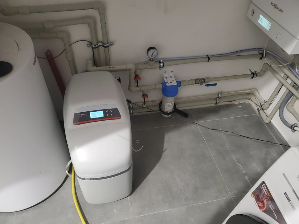 Biała stacja uzdatniania wody Viessmann z wyświetlaczem, obok zbiornik akumulacyjny i piec gazowy na ścianie, widoczne rury instalacji hydraulicznej i filtr wody z manometrem na szarej podłodze.