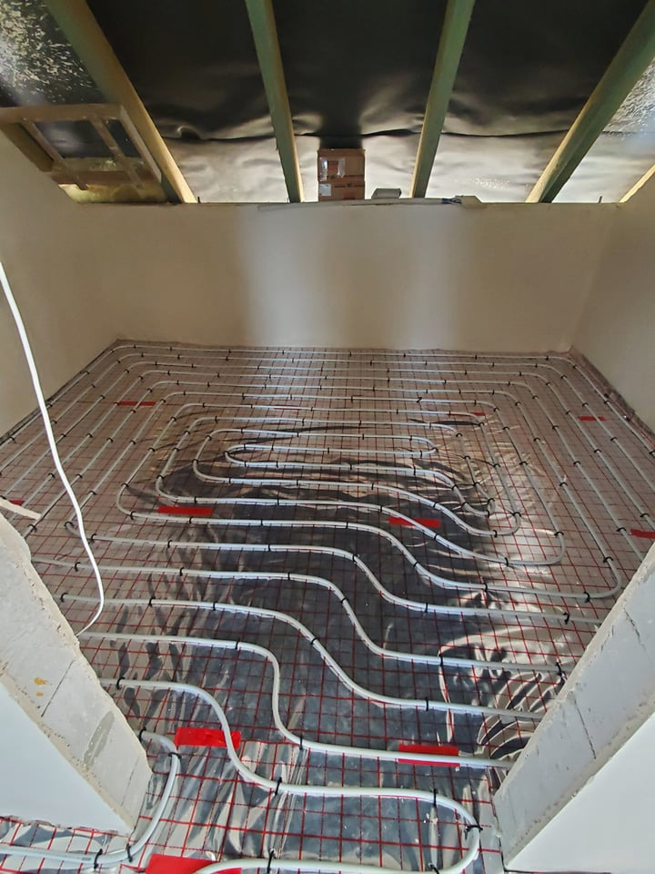 Montaż ogrzewania podłogowego: białe rurki ułożone na folii z czerwoną siatką w pomieszczeniu z surowym stropem i ścianami w trakcie wykończenia.