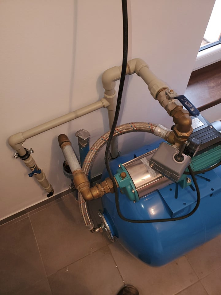 Niebieski hydrofor z podłączonymi rurami PCV i elastycznym przewodem w pomieszczeniu, widoczne nieszczelności i wyciek wody na podłodze z płytek.