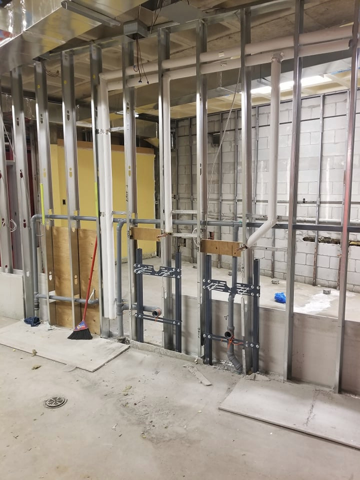 Surowe wnętrze w trakcie budowy z widocznymi metalowymi profilami ścian, instalacją wodno-kanalizacyjną z szarych rur, częściowo zamontowanymi uchwytami i fragmentem miotły opartą o ścianę.