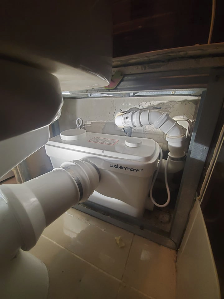 Biała przepompownia ścieków Waterman Pro, z podłączonymi rurami PCV, zamontowana w ciasnej przestrzeni w Konstancinie-Jeziornej. Widoczne betonowe podłoże i metalowa rama.