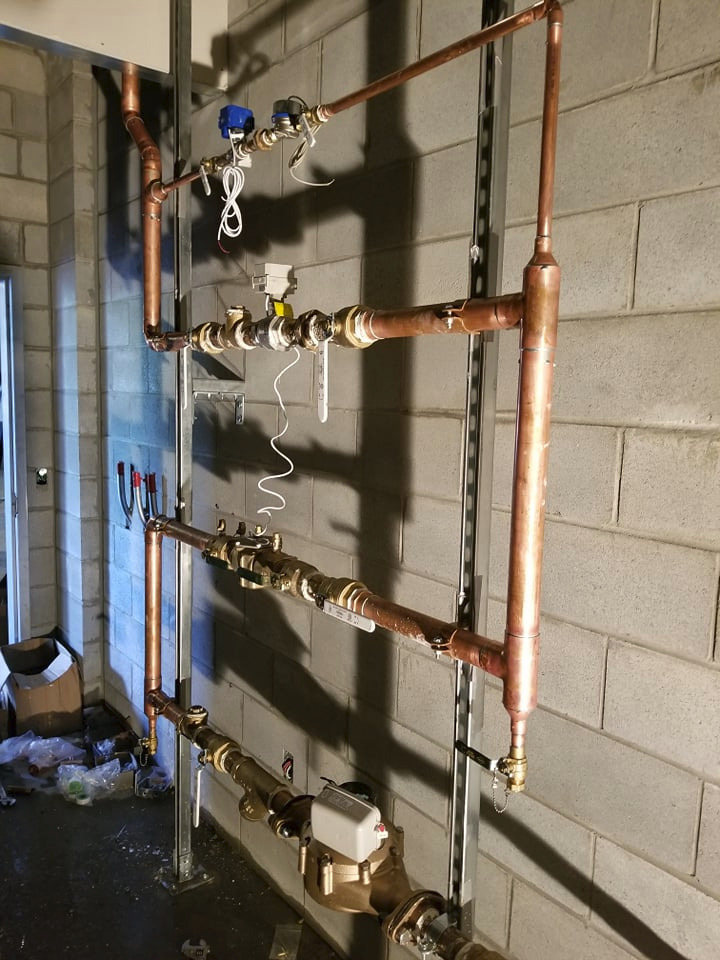 Miedziana instalacja wodna z zaworami i licznikiem, zamontowana na metalowej konstrukcji przy szarej, ceglanej ścianie w surowym wnętrzu.