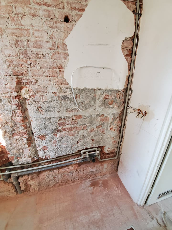 Fragment ściany z odsłoniętą czerwoną cegłą, częściowo pokrytą tynkiem, z widocznymi rurami instalacji wodnej i przewodami elektrycznymi. Ślady po kuciu w ścianie i na podłodze.