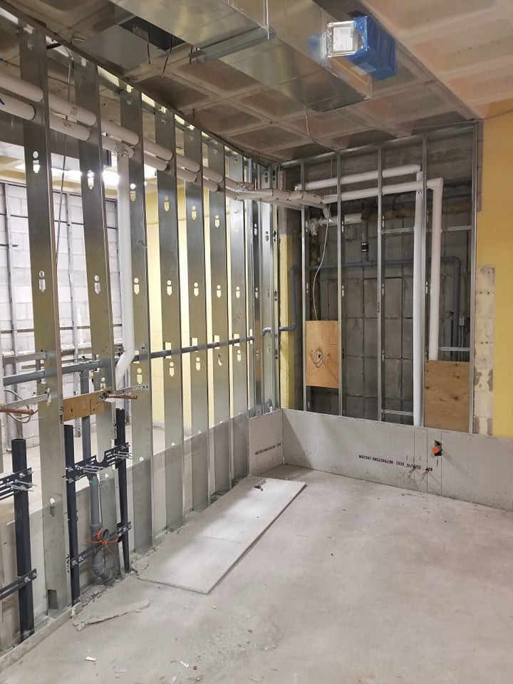 Wnętrze w trakcie instalacji wodno-kanalizacyjnej: widoczne rury, metalowe profile mocujące, częściowo wykończone ściany z płyt gipsowo-kartonowych i odsłonięte instalacje elektryczne.