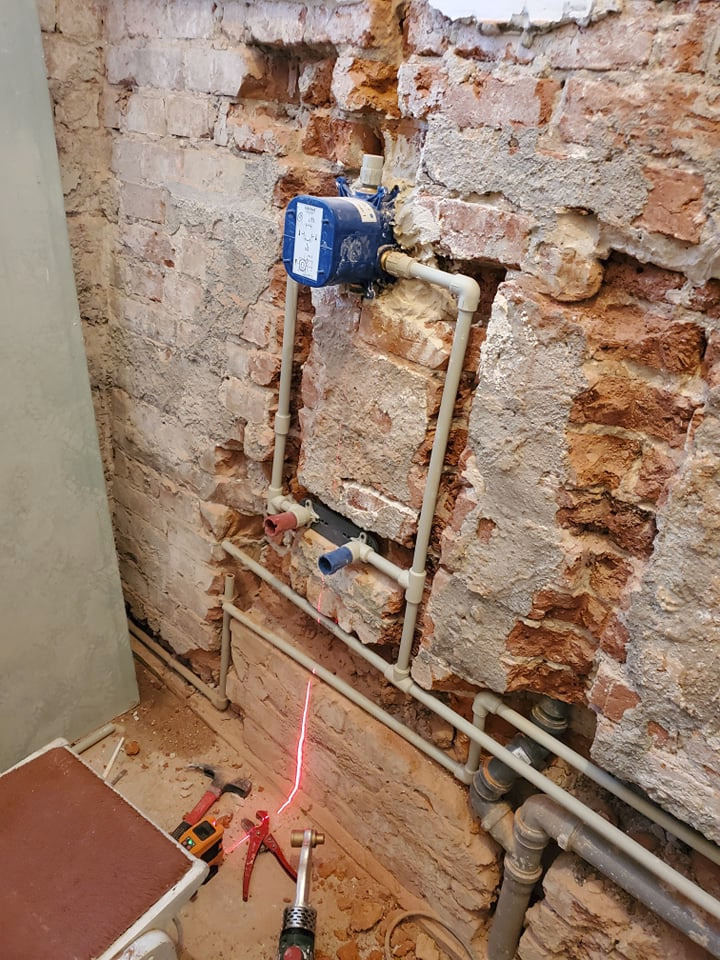 Instalacja wodna w trakcie montażu, widoczne rury PCV, niebieski zawór, laserowy poziom, narzędzia na podłodze, surowa ceglana ściana z ubytkami tynku.