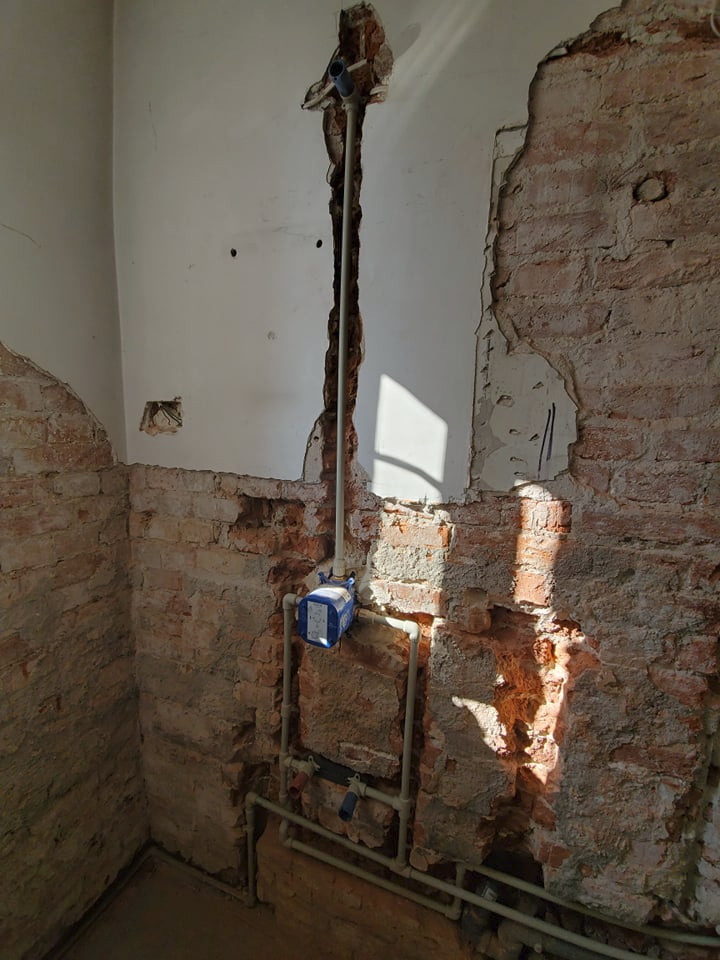 Ściana w trakcie remontu z odsłoniętą cegłą i nową instalacją wodną z niebieskim zaworem, pionowa rura w wyżłobieniu ściany.