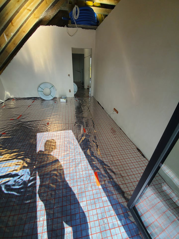 Poddasze w trakcie instalacji ogrzewania podłogowego; widoczna mata grzewcza z czerwonymi liniami, rury na ścianie, oraz cień osoby fotografującej.
