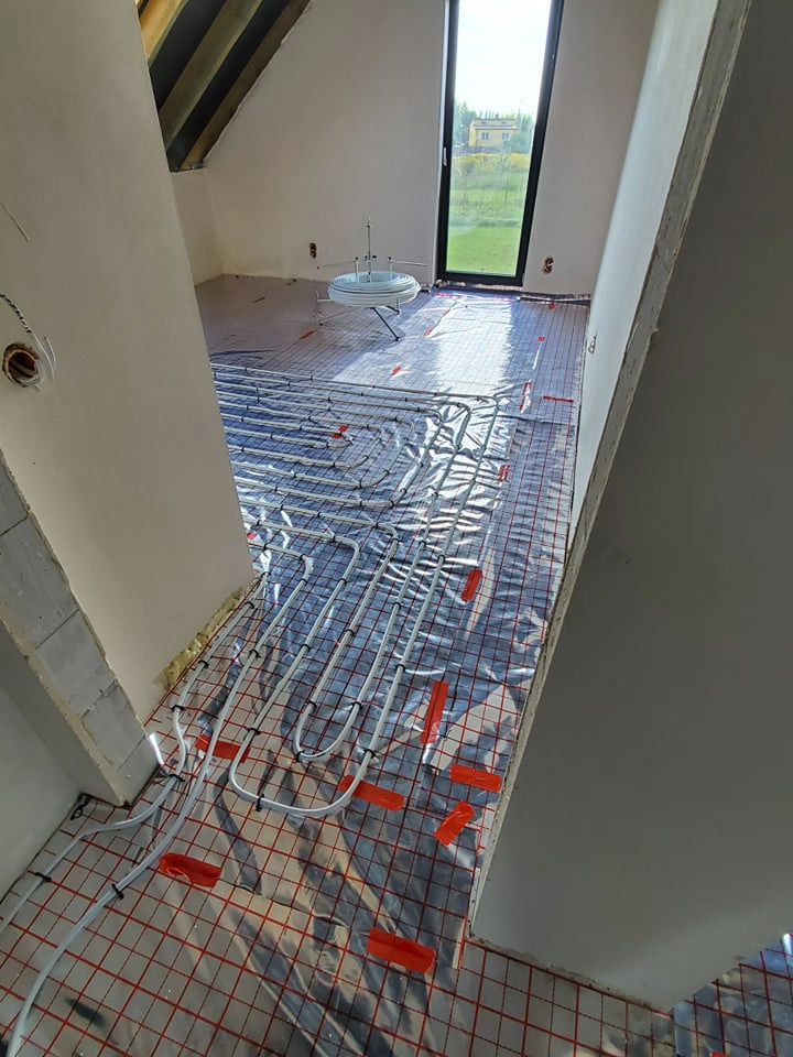 Białe rurki ogrzewania podłogowego ułożone na folii z czerwoną siatką w jasnym pomieszczeniu z oknem i widoczną konstrukcją dachu.