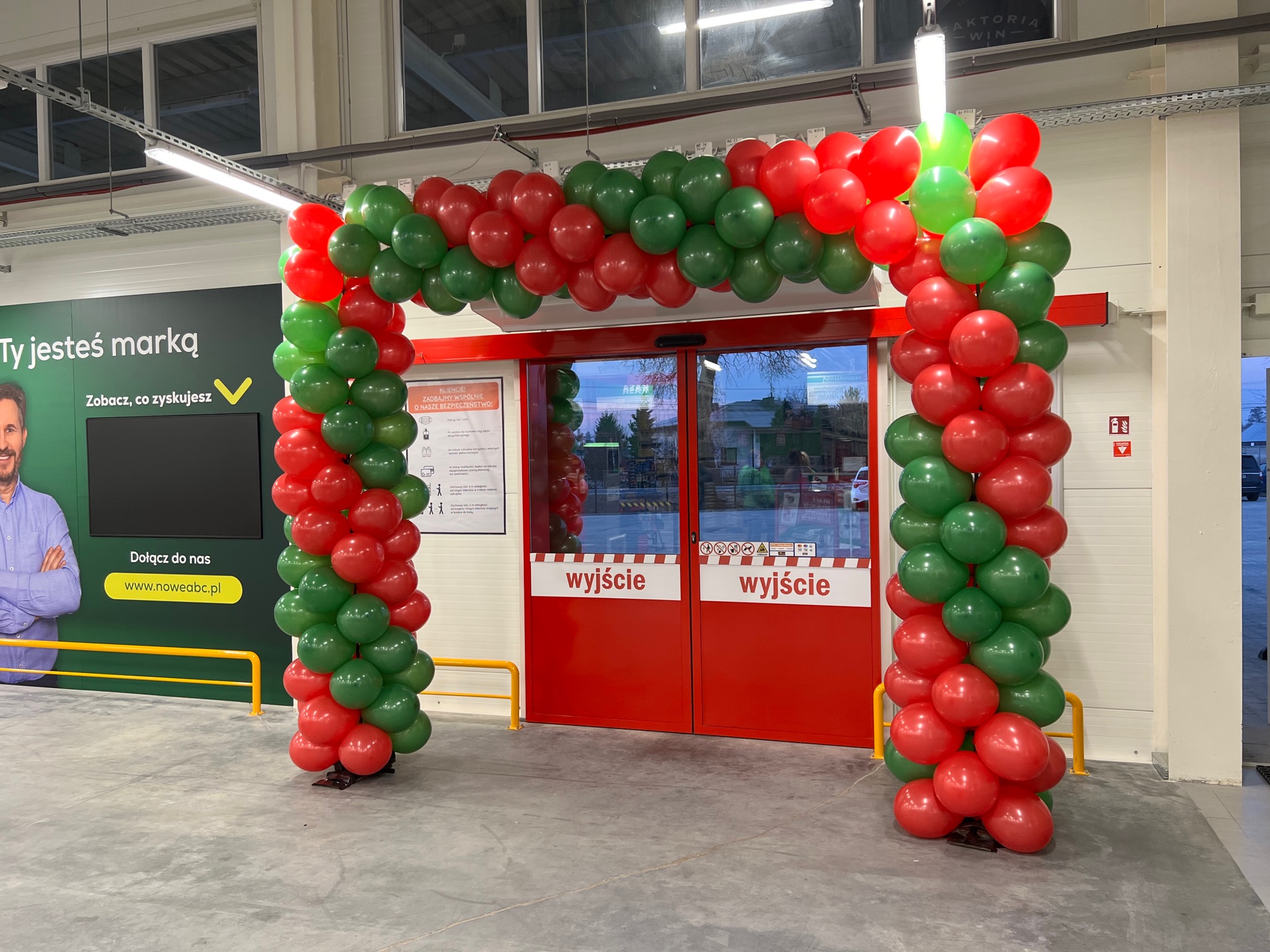 Dekoracja wejścia do sklepu balonami w kolorach czerwonym i zielonym, tworząca łuk nad drzwiami oznaczonymi jako wyjście.