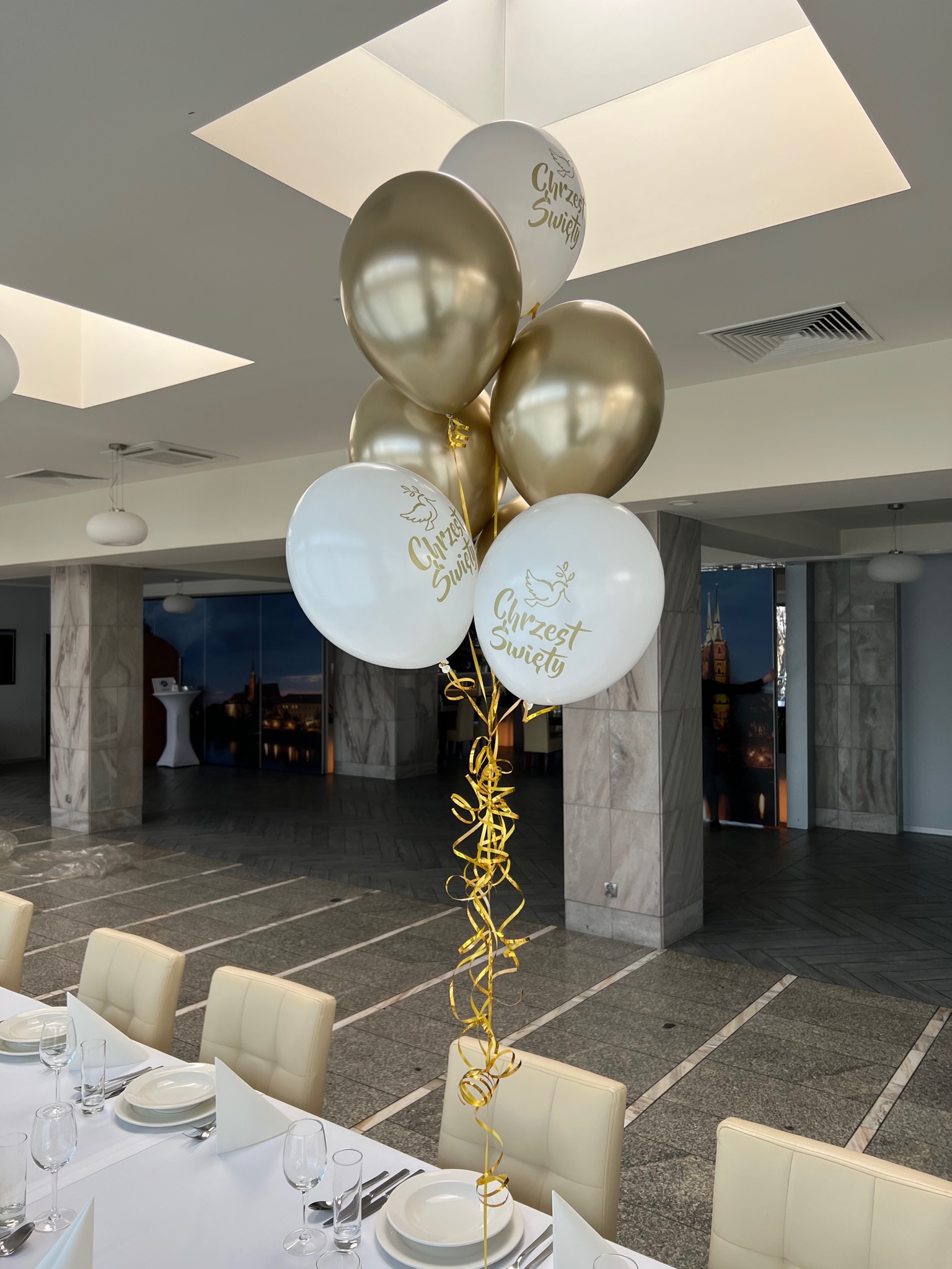 Dekoracja stołu na uroczystość Chrztu Świętego: białe balony z napisem i złotym gołąbkiem, złote balony, kręcone złote wstążki, nakryty stół z białym obrusem i zastawą.
