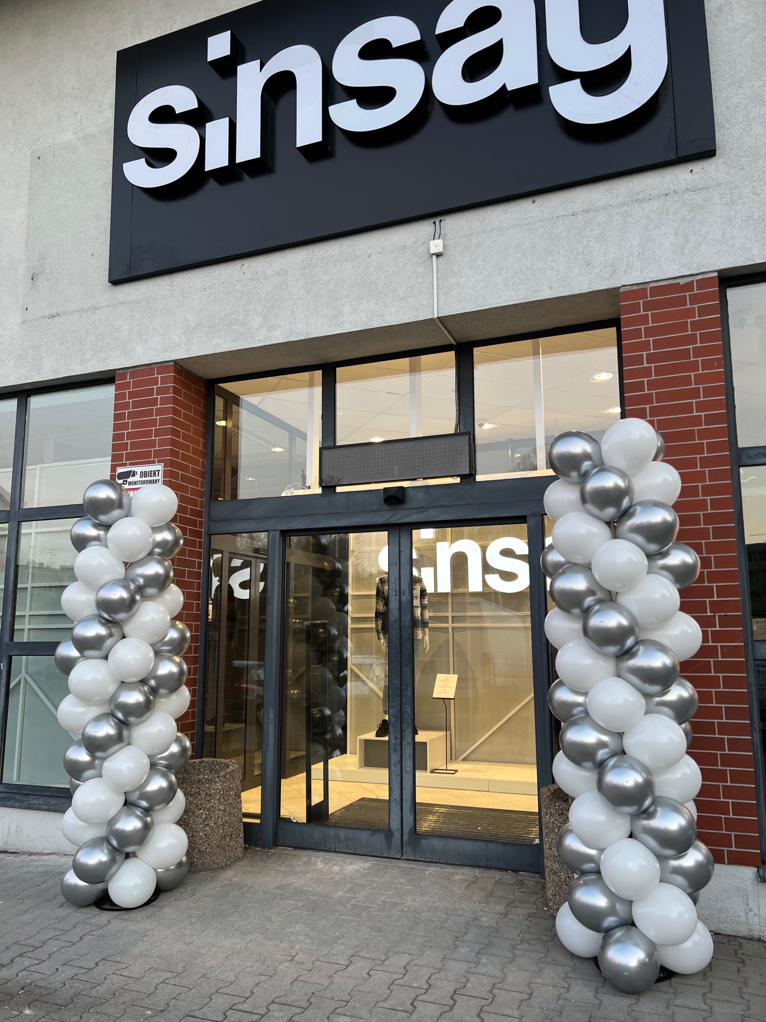 Wejście do sklepu udekorowane kolumnami z białych i srebrnych balonów; widoczny szyld z nazwą sklepu.