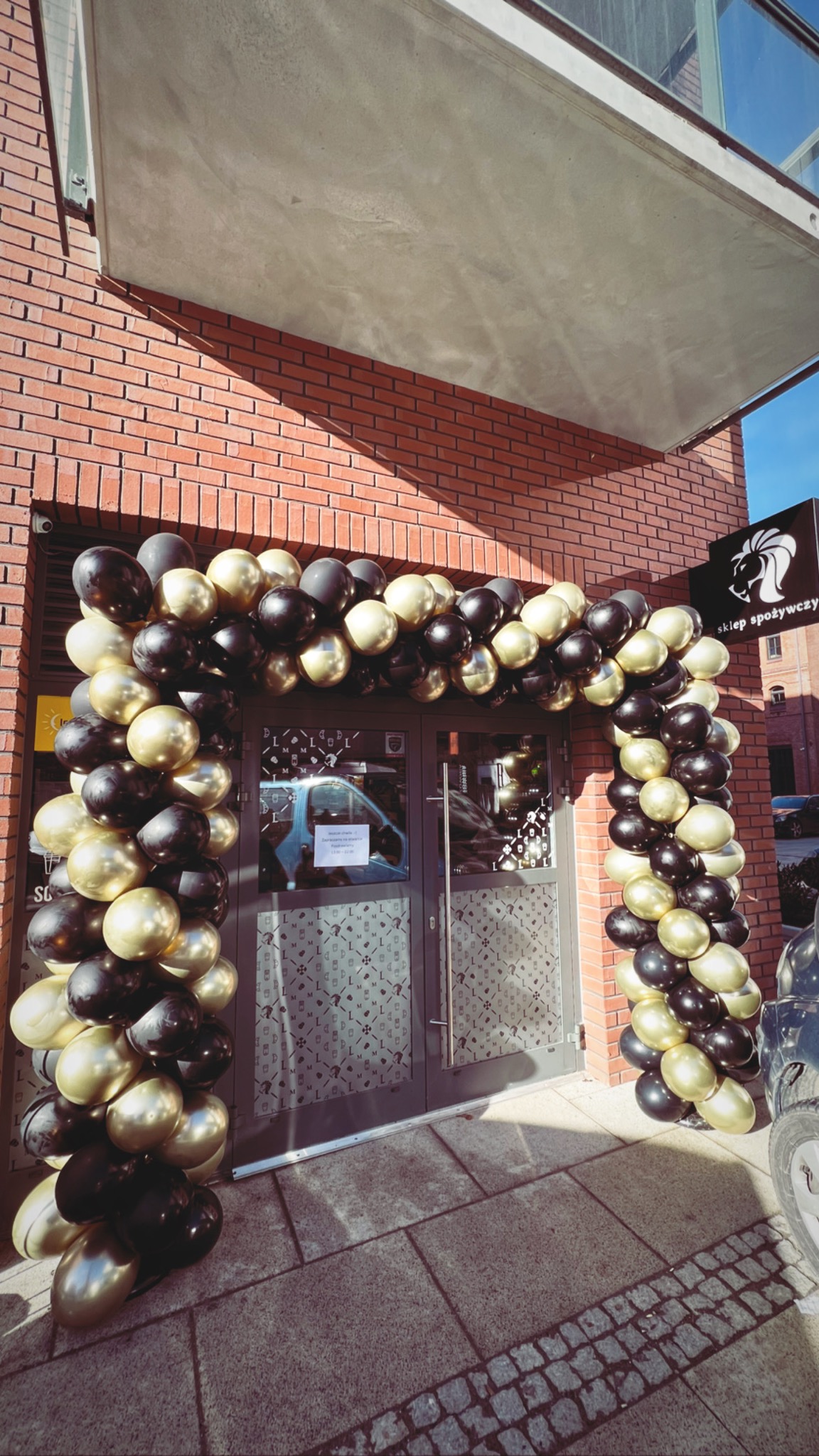 Wejście do sklepu spożywczego udekorowane łukiem z czarno-złotych balonów, ceglana fasada budynku, szyld z logo sklepu po prawej stronie.