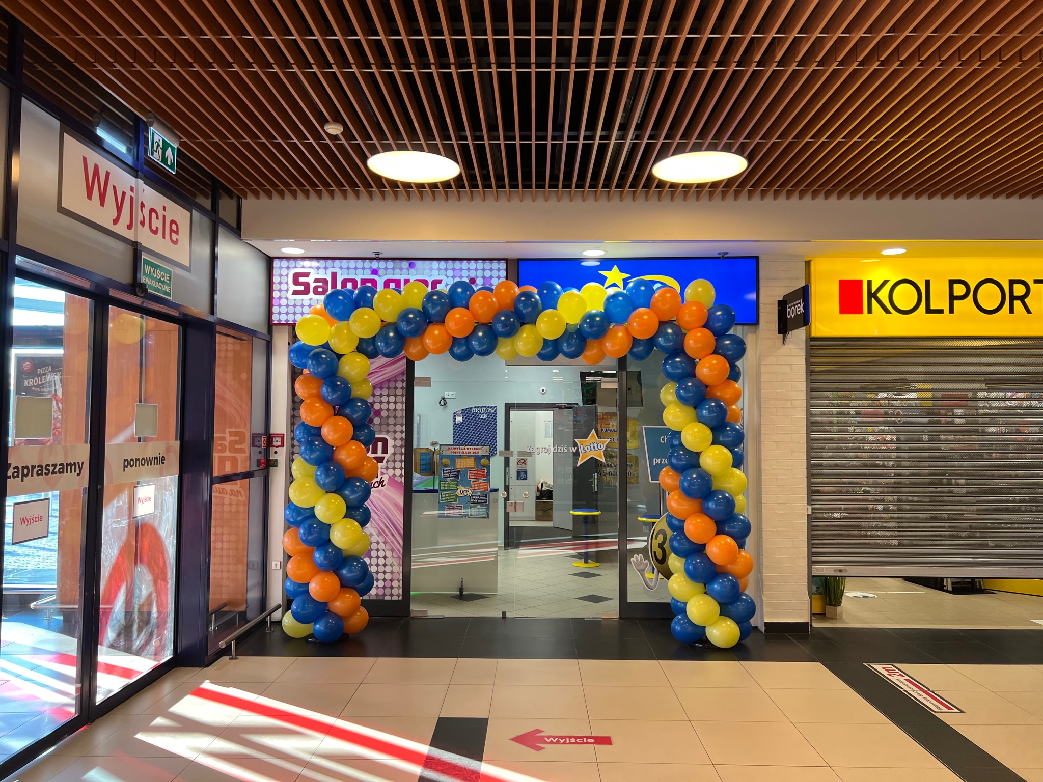 Dekoracja wejścia do salonu balonami w kolorach niebieskim, pomarańczowym i żółtym, w tle widoczny lokal z napisem Lotto i Kolport, sufit z drewnianych listew.