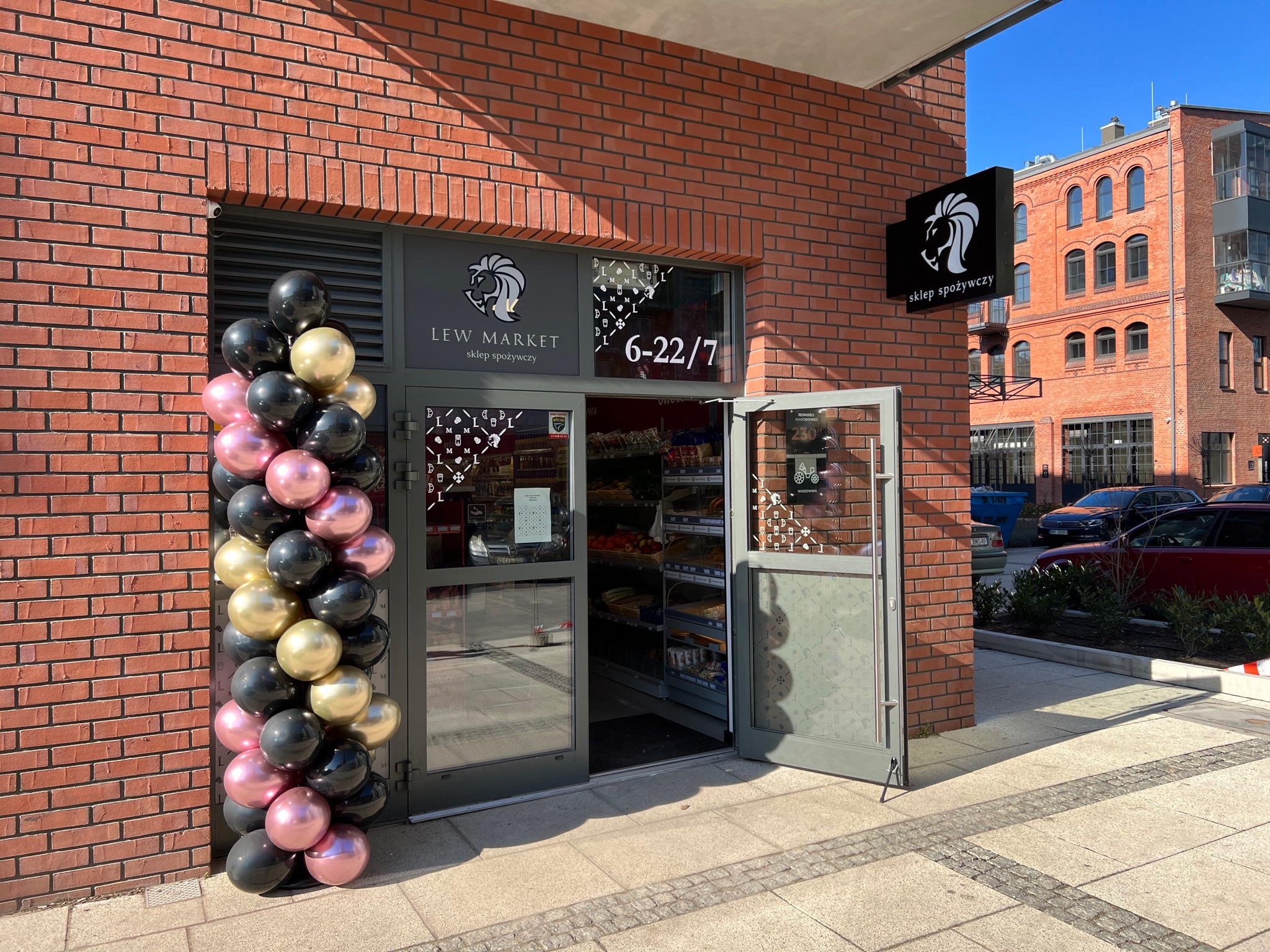 Wejście do sklepu spożywczego Lew Market udekorowane girlandą z czarnych, złotych i różowych balonów, widoczny szyld z logo lwa i godziny otwarcia.