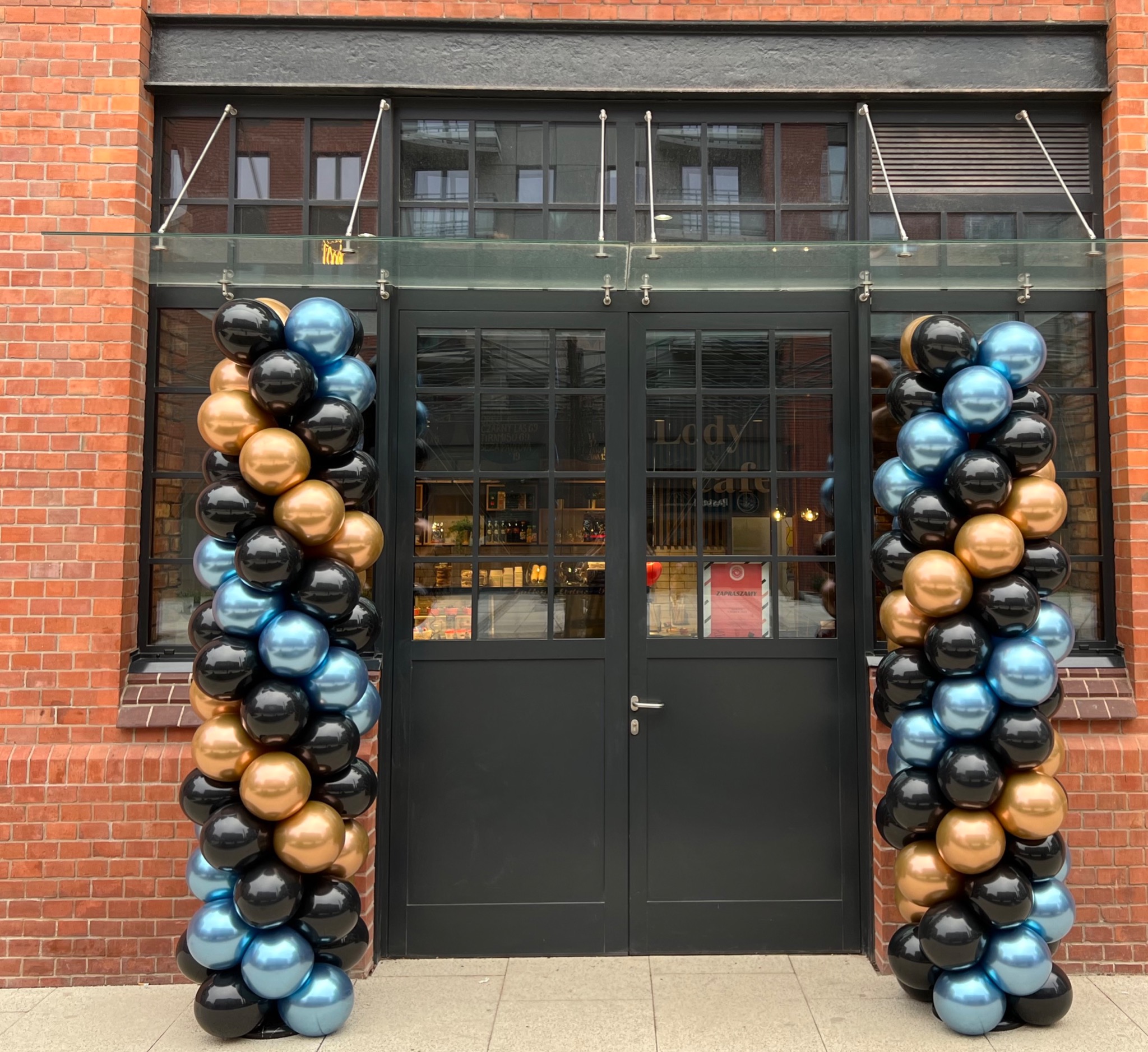 Wejście do lokalu ozdobione kolumnami z balonów w kolorach: czarnym, złotym i niebieskim. Balony ułożone pionowo po obu stronach ciemnych, przeszklonych drzwi w budynku z czerwonej cegły.