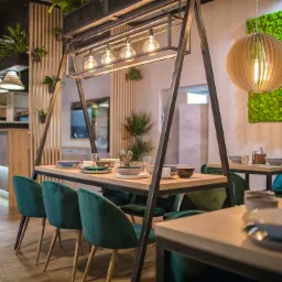 Nowoczesna aranżacja wnętrza restauracji z drewnianymi elementami, zieloną ścianą z mchu, metalową konstrukcją nad stołem z oświetleniem i zielonymi aksamitnymi krzesłami.