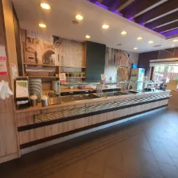 Wnętrze samoobsługowej restauracji z bufetem, widoczny ciąg zabudowy meblowej z beżowego drewna z metalowymi elementami, za nim ściana z fototapetą imitującą kamienne miasto, oświetlenie sufitowe...