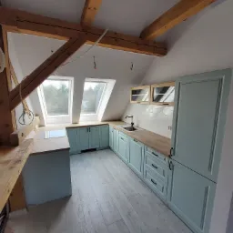 Jasna kuchnia w skosach poddasza z drewnianymi belkami stropowymi, wyposażona w błękitne szafki z frezowanymi frontami i drewniane blaty, widok na okna dachowe.