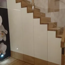 Schody drewniane z zabudową szaf na wymiar z białymi frontami, przeszklona balustrada, oświetlenie punktowe pod schodami