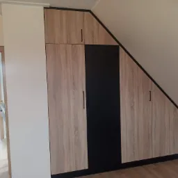 Szafa wnękowa z frontami w kolorze jasnego drewna i czarną wstawką, dopasowana do skosu poddasza, z widocznymi gniazdkami elektrycznymi na ścianie.