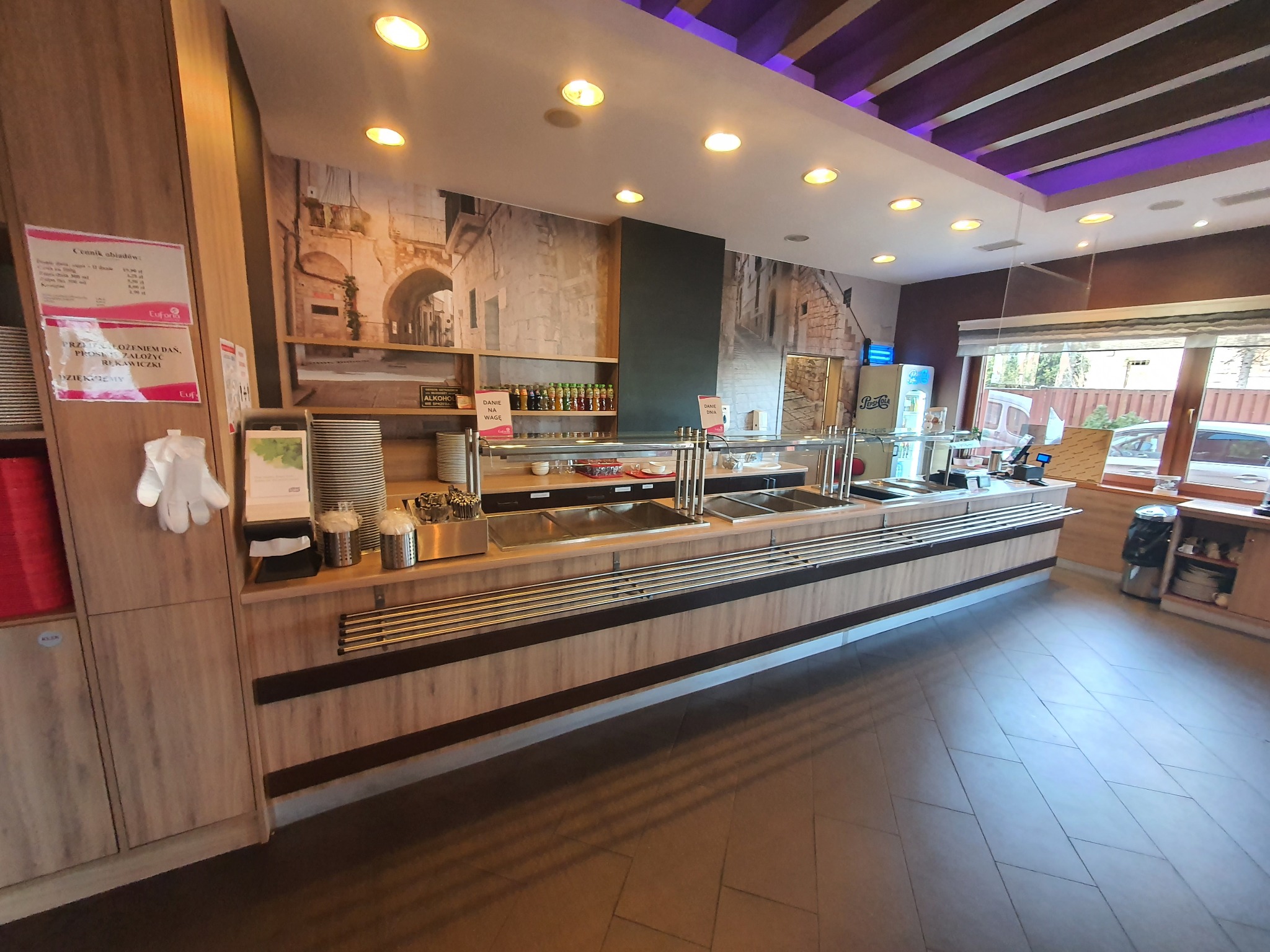 Wnętrze samoobsługowej restauracji z bufetem, widoczny ciąg zabudowy meblowej z beżowego drewna z metalowymi elementami, za nim ściana z fototapetą imitującą kamienne miasto, oświetlenie sufitowe...