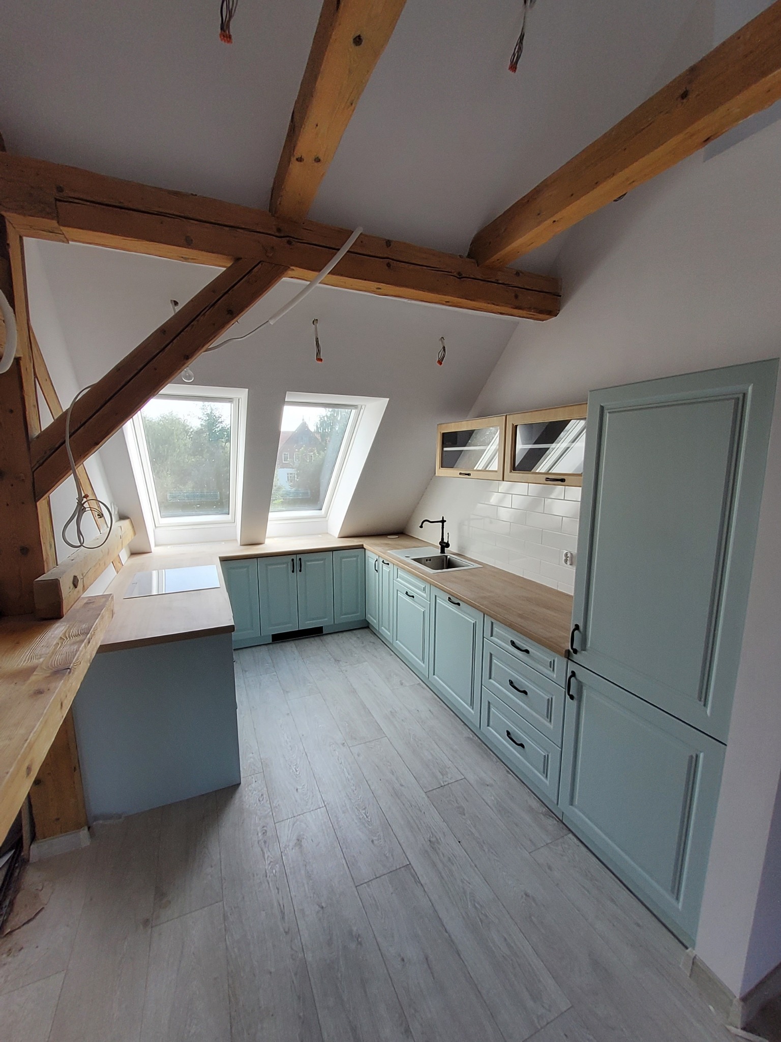 Jasna kuchnia w skosach poddasza z drewnianymi belkami stropowymi, wyposażona w błękitne szafki z frezowanymi frontami i drewniane blaty, widok na okna dachowe.