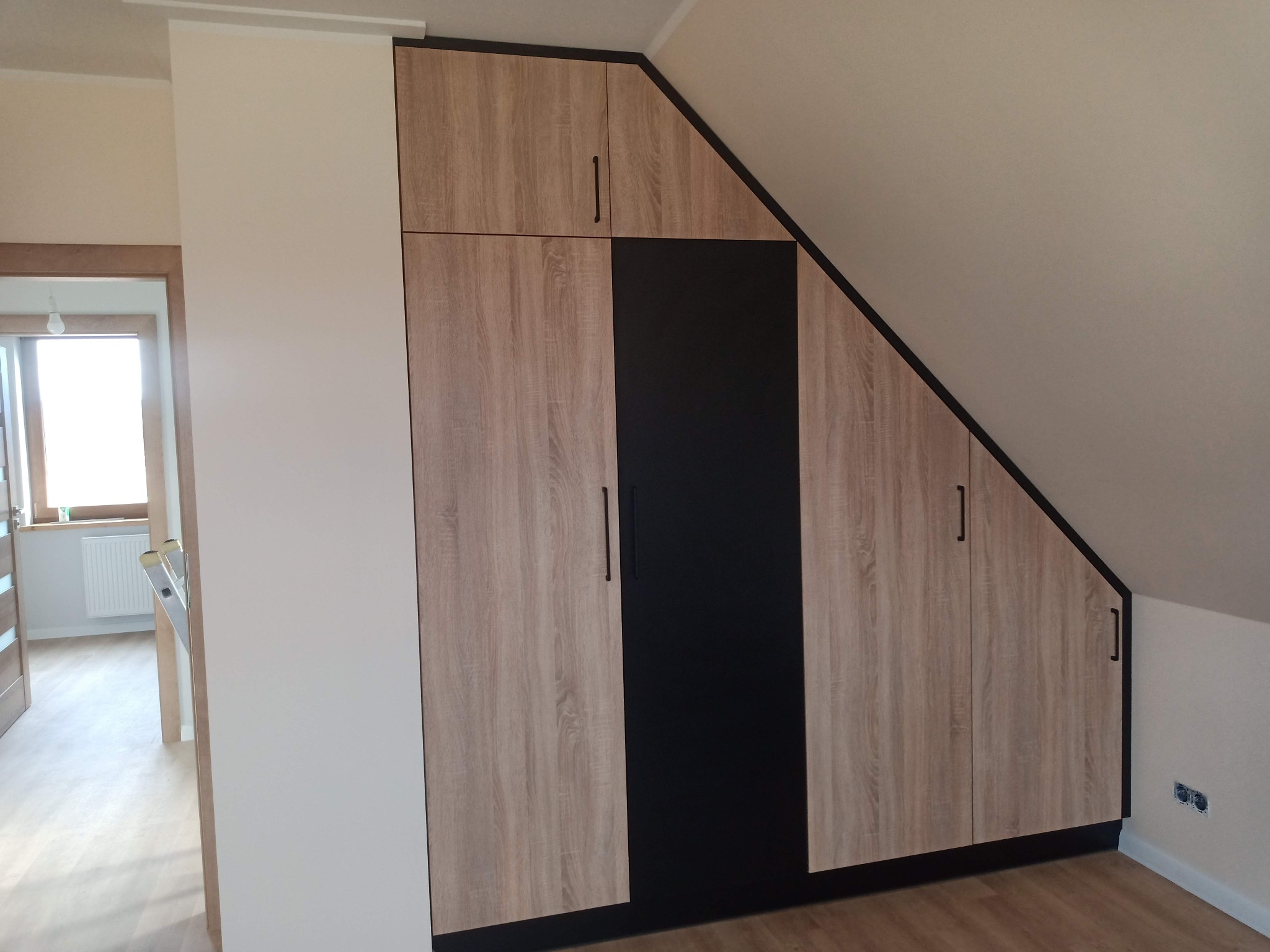 Szafa wnękowa z frontami w kolorze jasnego drewna i czarną wstawką, dopasowana do skosu poddasza, z widocznymi gniazdkami elektrycznymi na ścianie.