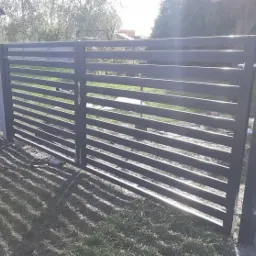 Nowoczesna, pozioma, metalowa brama wjazdowa w kolorze grafitowym, zamontowana pomiędzy betonowym słupkiem a metalowym słupem, na tle trawnika i drzew