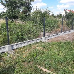 P.P.H.U. MM MAŁGORZATA KITA - Nowo postawione ogrodzenie z siatki z podmurówką betonową w jasnoszarym odcieniu, z czarnymi słupkami. Trawa rosnąca przy ogrodzeniu.