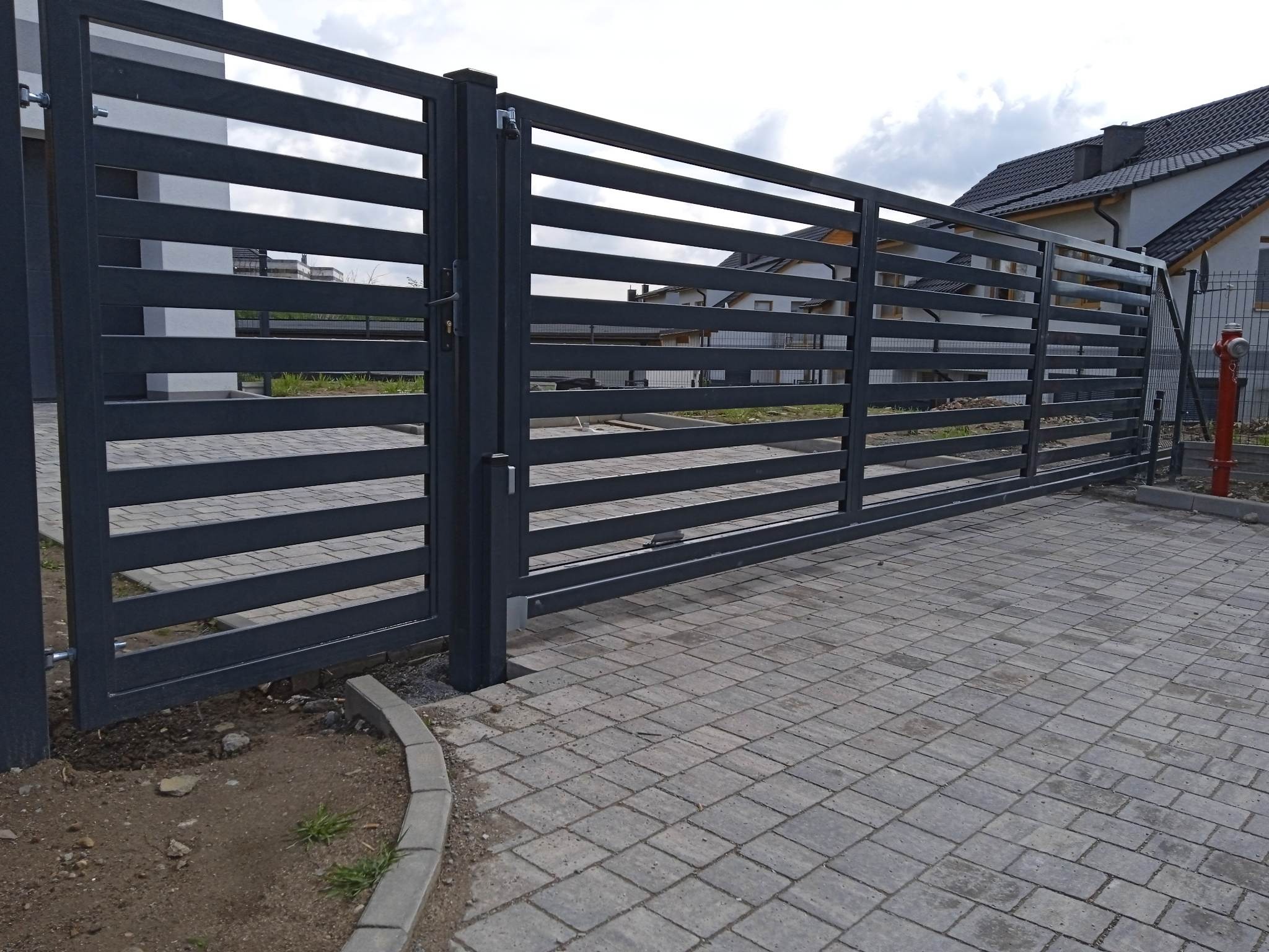 Nowoczesna, przesuwna brama wjazdowa z poziomymi, szarymi panelami metalowymi i widoczną kostką brukową na podjeździe, na tle domu.