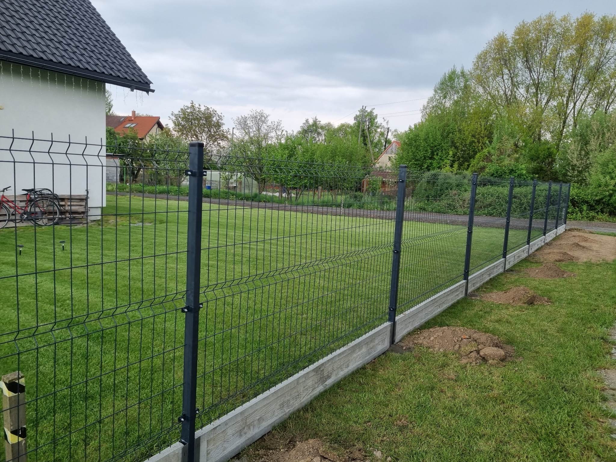 Ciemna siatka panelowa zamontowana na betonowej podmurówce, widok na trawnik i dom w tle. Świeżo usypana ziemia przy ogrodzeniu.