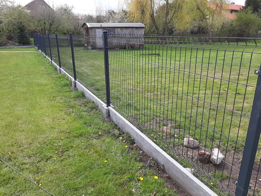 Nowoczesne ogrodzenie panelowe z podmurówką betonową na zielonym terenie, w tle drewniana altana. Widok z niskiej perspektywy podkreśla linię ogrodzenia.