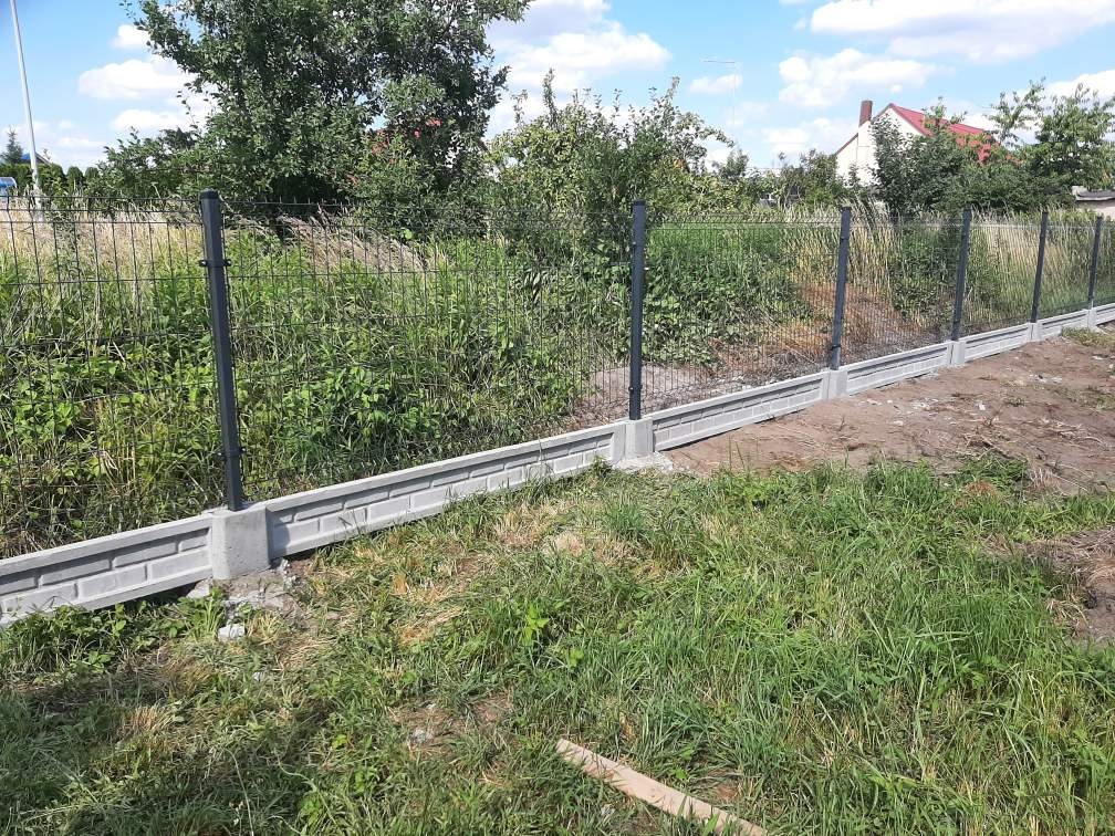 Nowo postawione ogrodzenie z siatki z podmurówką betonową w jasnoszarym odcieniu, z czarnymi słupkami. Trawa rosnąca przy ogrodzeniu.