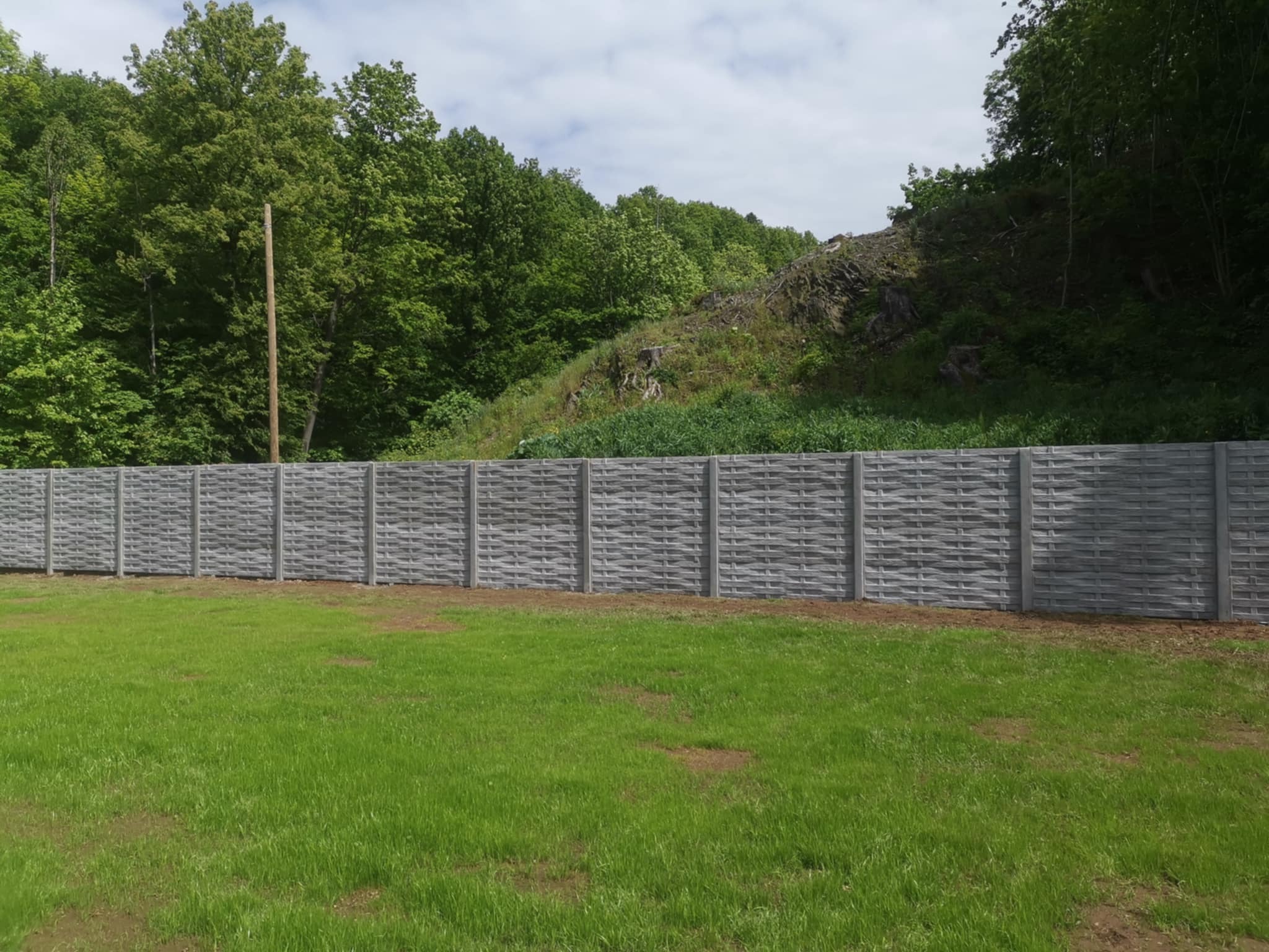 Długi, szary, betonowy płot o wzorze plecionki, oddzielający zielony trawnik od zalesionego terenu. W tle widoczny słup energetyczny.
