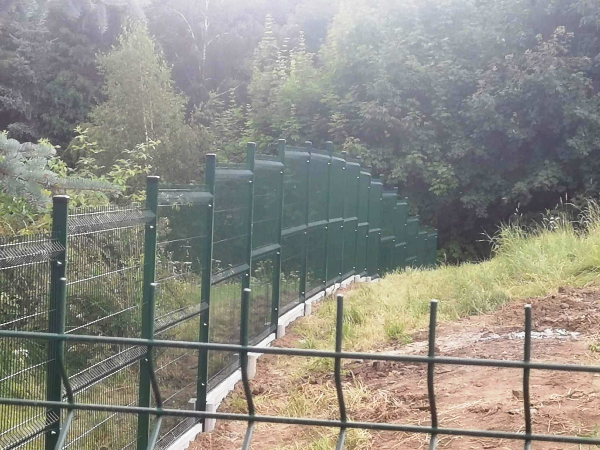 Zielone ogrodzenie panelowe z podmurówką na nierównym terenie, widoczne słupki i siatka, w tle zieleń drzew, ujęcie z dołu.