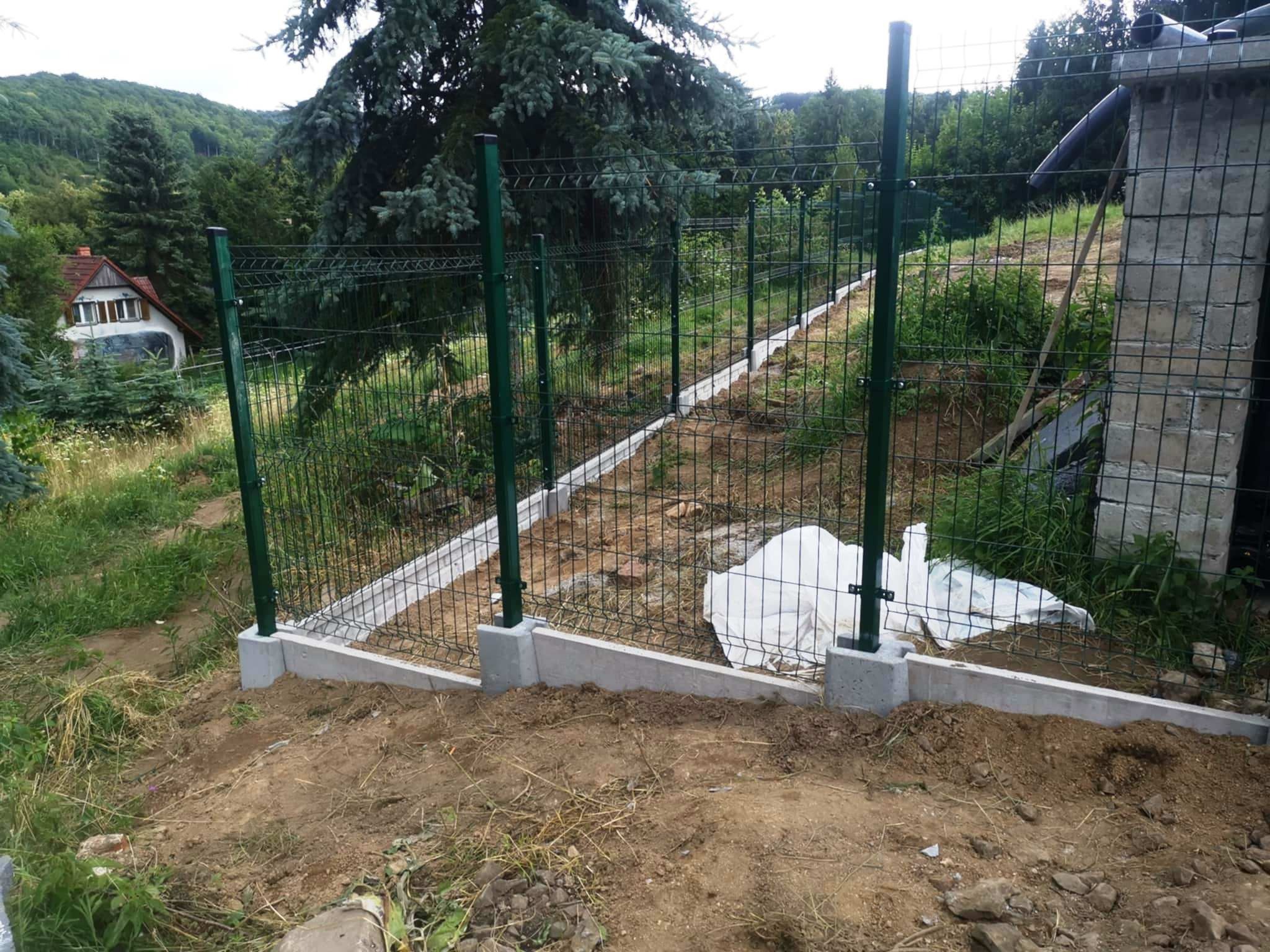 Montaż ogrodzenia panelowego z zielonymi słupkami na betonowej podmurówce, widok na pagórkowaty teren i dom w tle. Częściowo rozłożona folia budowlana.