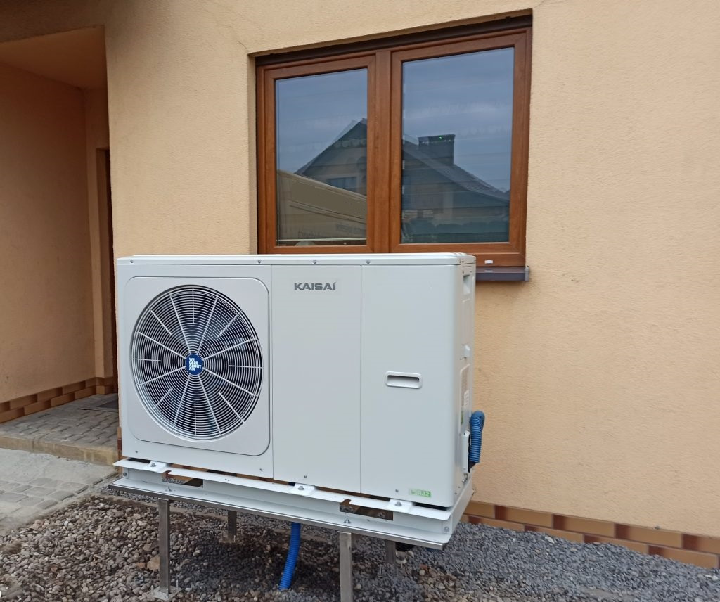 Zewnętrzna jednostka pompy ciepła Kaisai, zamontowana na metalowej konstrukcji przy ścianie budynku w Krakowie, widoczna brązowa rama okienna w tle.