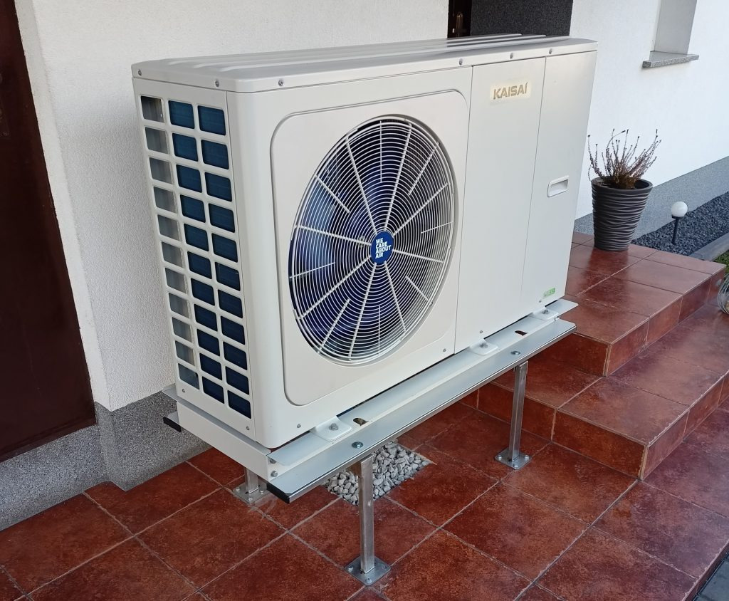 Zewnętrzna jednostka pompy ciepła Kaisai, zamontowana na metalowej konstrukcji przy schodach wejściowych do domu, widok z boku.