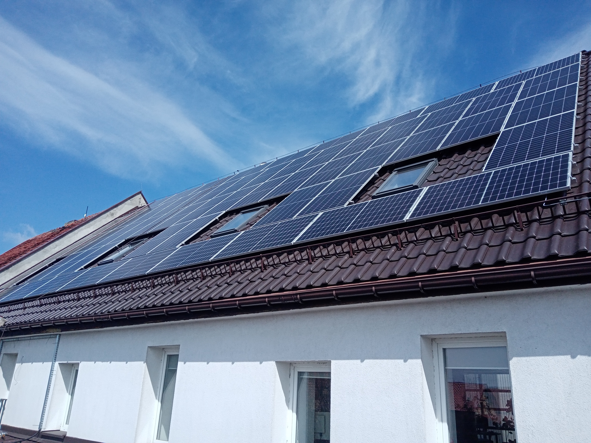 Panele fotowoltaiczne zamontowane na brązowym dachu domu z oknami dachowymi, widok z dołu na tle błękitnego nieba z chmurami.