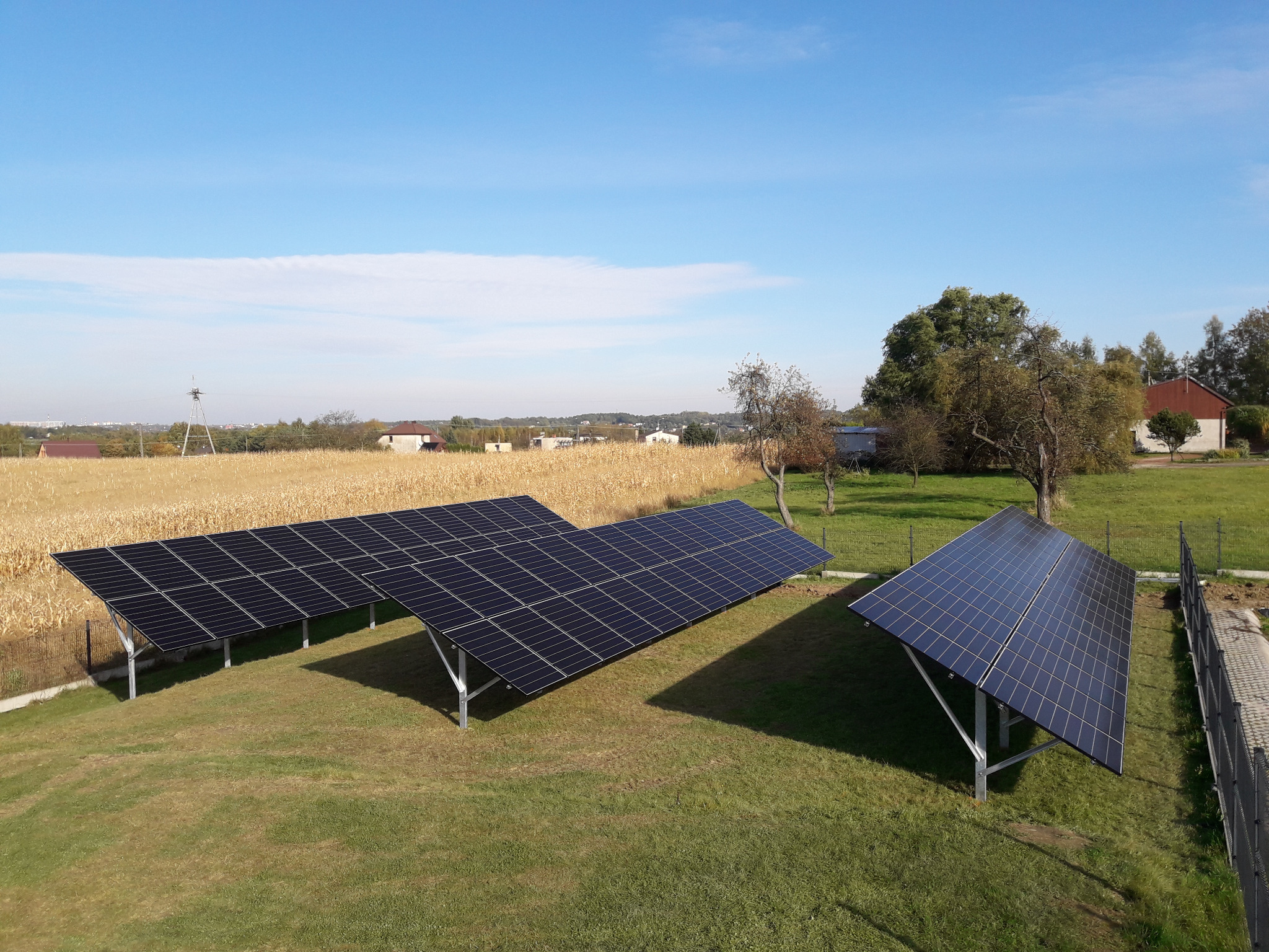 Instalacja fotowoltaiczna na trawiastym terenie z widokiem na pole zboża i zabudowania w oddali pod błękitnym niebem.