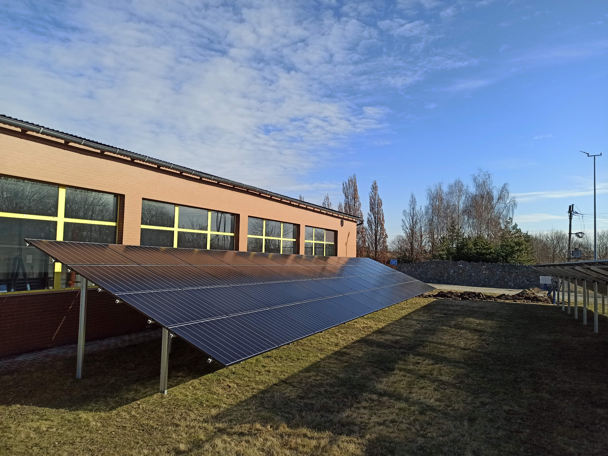 Rząd paneli słonecznych zamontowanych na metalowej konstrukcji przed budynkiem z cegły z żółtymi ramami okiennymi, na tle błękitnego nieba z chmurami.