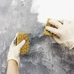 Ręce w lateksowych rękawicach nanoszą gąbką szarą farbę na białą ścianę, tworząc efekt dekoracyjny.