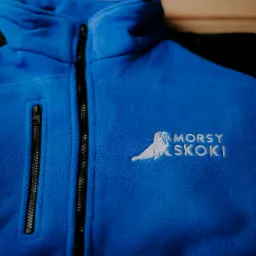 Niebieska polarowa bluza z czarnymi rękawami i białym haftowanym logo 'Morsy Skoki' z wizerunkiem morsa, leżąca na drewnianym stole.