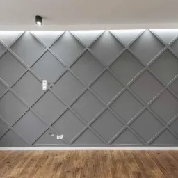Szara ściana z dekoracyjnymi listwami tworzącymi geometryczny wzór rombów, oświetlona od góry listwą LED, z widocznymi gniazdkami elektrycznymi i włącznikiem światła, oraz podłogą z paneli...