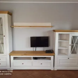 Zestaw mebli do salonu w stylu rustykalnym, składający się z dwóch białych witryn z drewnianymi blatami i przeszklonymi drzwiczkami, połączonych szafką RTV z szufladami i półkami, na ścianie...