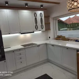 Jasna kuchnia z białymi szafkami, czarnym piekarnikiem i marmurową podłogą, widok na zewnątrz przez duże okno, ozdobna lampa wisząca.