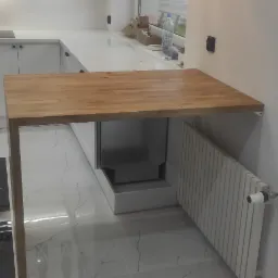 Drewniany blat przymocowany do ściany w nowoczesnej kuchni z białymi szafkami i czarno-białą podłogą, widok na ogród przez okno.