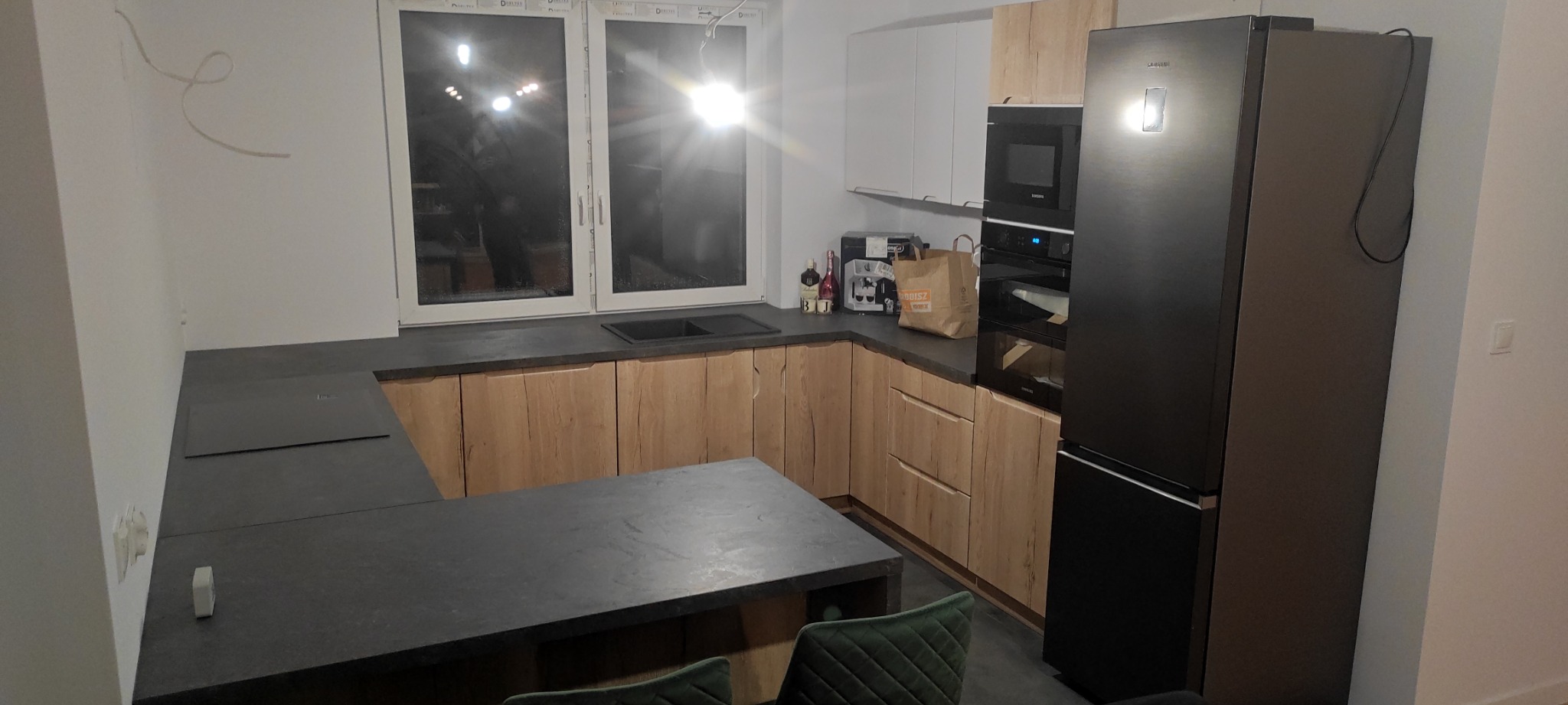 Nowoczesna kuchnia z drewnianymi szafkami o wyraźnej fakturze, czarnym blatem i sprzętem AGD w kolorze czarnym, uzupełniona o zielone krzesła barowe i widok na okno z intensywnym światłem.