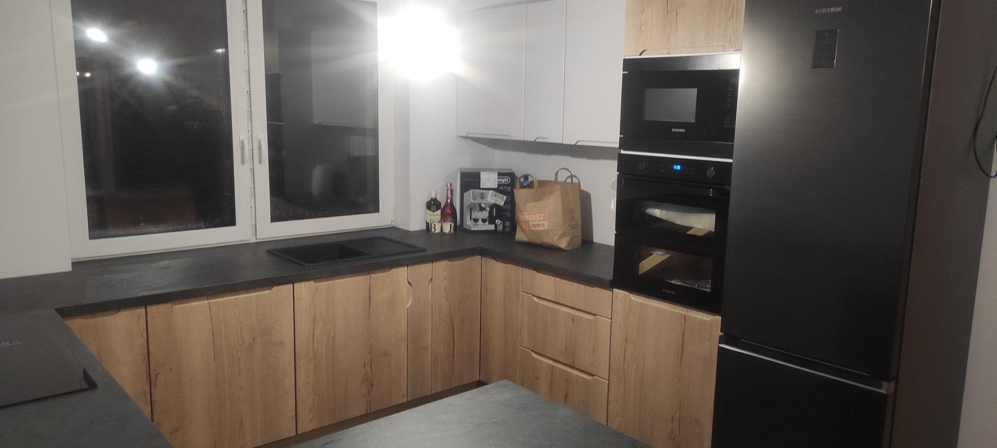 Nowoczesna kuchnia w odcieniach drewna i szarości, z czarnym zlewem pod oknem i zabudowaną czarną lodówką Samsung, oświetlona światłem dziennym